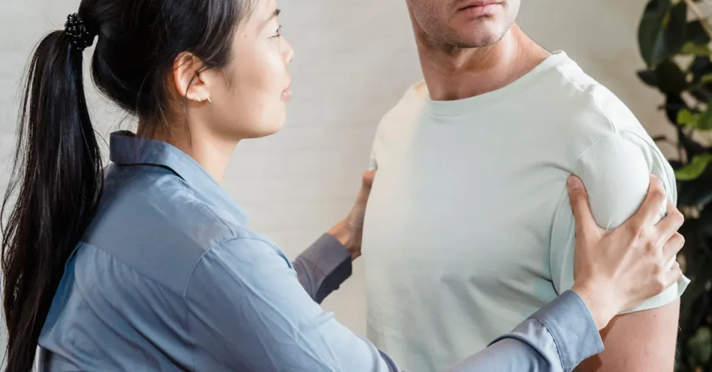 Woman in a blue shirt holds a man's shoulders, guiding him in a posture check, in a bright room.