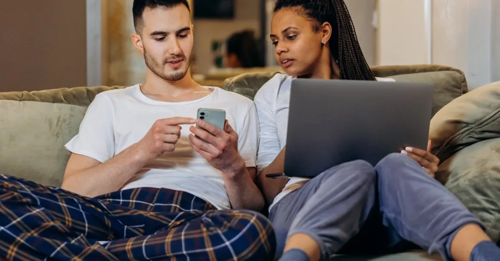 Couple sitting on a couch, the man points at a smartphone while the woman holds a laptop beside him in a living room setting.