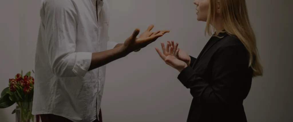 Two professionals converse, using expressive hand gestures, with a vase of red flowers in the background on the left.
