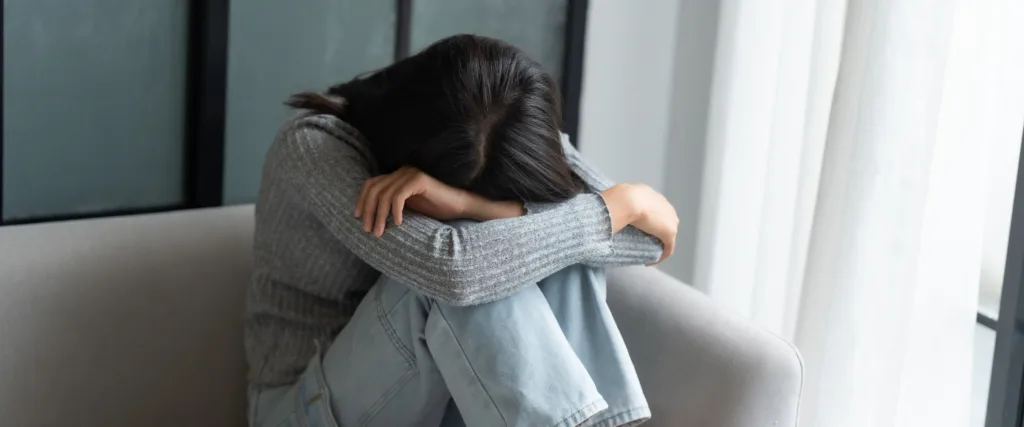 Person sitting on a sofa with head bowed and arms wrapped over their knees, looking distressed or overwhelmed.
