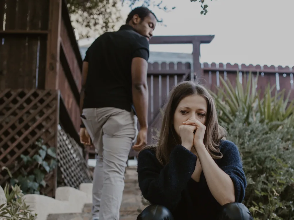 Woman crying while sitting, mascara dripping down her eyes. A man behind her wakling away while looking back.